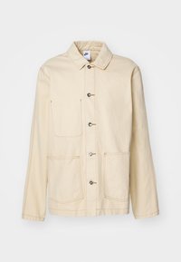 Beige jacka med knäppning fram, två bröstfickor och sidfickor, med kontrasterande sömmar och krage.