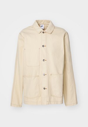 Beige jacka med knäppning fram, två bröstfickor och sidfickor, med kontrasterande sömmar och krage.
