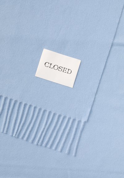 Lys blå tørklæde lavet af blødt materiale med frynser i kanten, ledsaget af en hvid mærkat, der står "CLOSED" med fede sorte bogstaver.