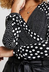 Schwarze Bluse mit unterschiedlich großen weißen Punkten. Sie hat ein Wickeldesign und Puffärmel und besteht aus einem glatten Stoff mit einer leicht glänzenden Textur.