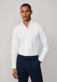 White long-sleeve button-up shirt with a subtle logo. Worn with dark navy pants. Features a classic collar and a smooth fabric texture.