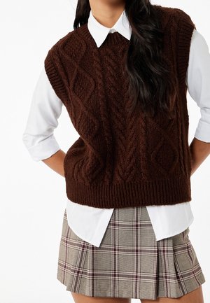 Femme portant un pull sans manches en maille torsadée marron foncé sur une chemise blanche à col et une jupe plissée à carreaux.