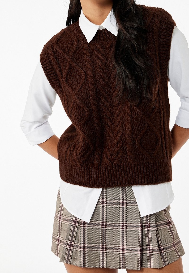 Femme portant un pull sans manches en maille torsadée marron foncé sur une chemise blanche à col et une jupe plissée à carreaux.