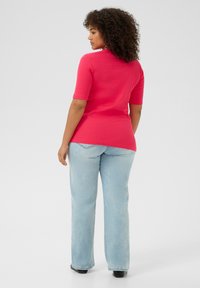 Top rosa ajustado y acanalado con mangas cortas y cuello alto, combinado con vaqueros azul claro y zapatos negros, visto desde atrás.