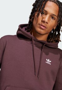 Mørkerød hettegenser laget av bomullsmiks, med en frontlomme, snorhette og hvit Adidas-logo på venstre bryst. Myk tekstur.