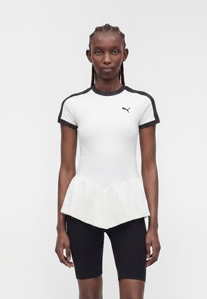 Frau mit geflochtenem Haar, die ein weißes, eng anliegendes Puma-Shirt mit schwarzem Besatz und Peplum-Saum trägt, kombiniert mit schwarzen Radlerhosen vor einfarbigem Hintergrund.