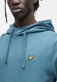 Kapuce pieguļošs džemperis zaļgani zili zaļā krāsā ar reljefu tekstūru. Kreisajā krūtīs izšūta zelta un melna logo, regulējamas auklas.