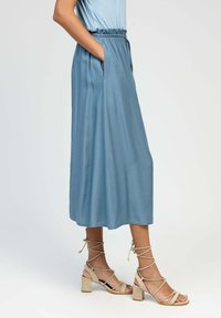 Une longue jupe fluide de couleur bleu clair avec une taille froncée et des poches latérales, associée à des sandales à talons blocs beige avec des lanières.