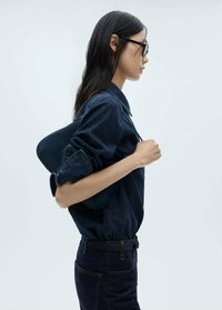 Dark denim jacket with quilted texture, paired with dark jeans. Model holding a dark bag, wearing black sunglasses, facing right.