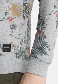 Jersey gris con patrones florales en colores apagados, puños acanalados y una etiqueta de marca en el dobladillo. Tela suave con un ajuste regular.