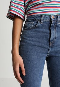 Jean en denim bleu taille haute, au design ajusté, avec des poches avant et une fermeture à bouton visible, porté avec un haut à rayures à manches courtes.