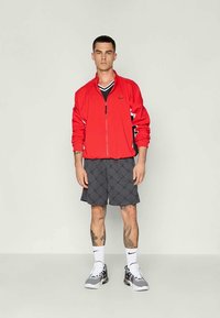 Raudona zip-up striukė su juodais akcentais, juodai balta V formos kaklo marškinėliai, pilki šortai su deimanto raštu, balti kojinės ir pilni sportiniai batai.