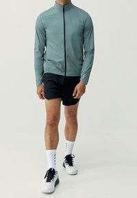 Lättvikts jacka i teal med hög krage och dragkedja framtill, tillsammans med svarta shorts. Modell som bär vita strumpor och svarta träningsskor.