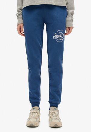 Pantalones de chándal azules hechos de una mezcla de algodón, con un logo impreso en el frente y puños acanalados en los tobillos. Combinados con zapatillas en tonos neutros.