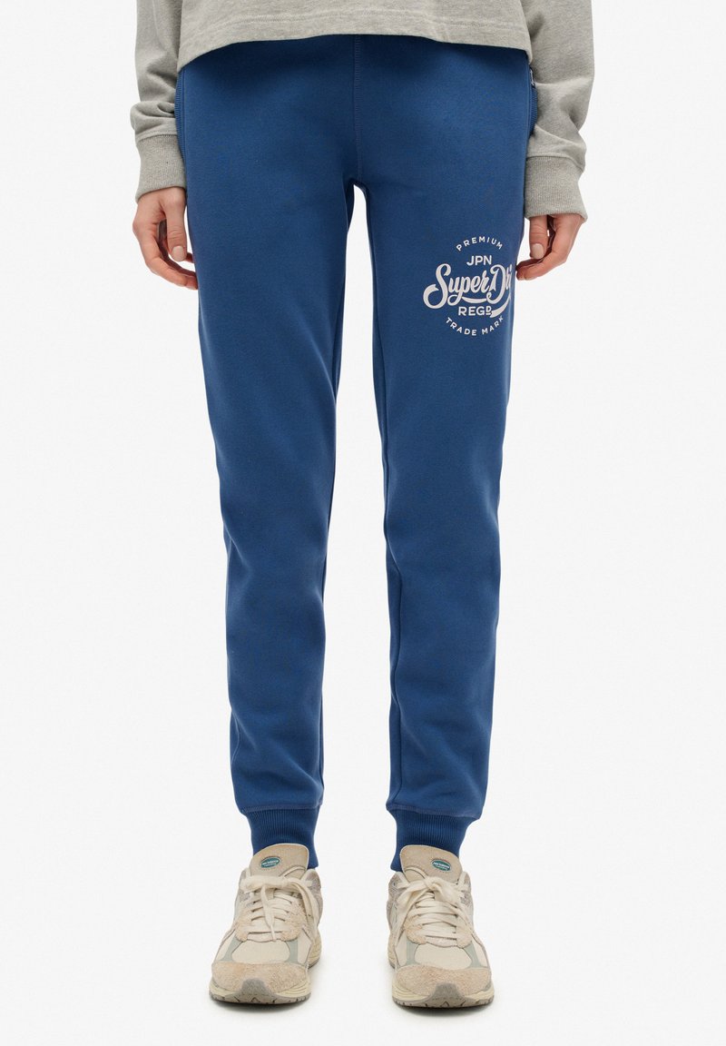 Niebieskie spodnie dresowe z mieszanki bawełny, z nadrukiem logo z przodu i ściągaczami na kostkach. W połączeniu z neutralnymi trampkami.