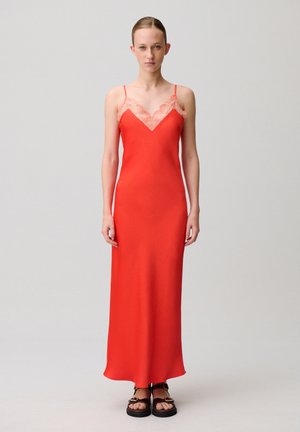 Donna in piedi che indossa un lungo abito sottoveste rosso con rifiniture in pizzo e sandali neri piatti, su uno sfondo grigio chiaro uniforme.