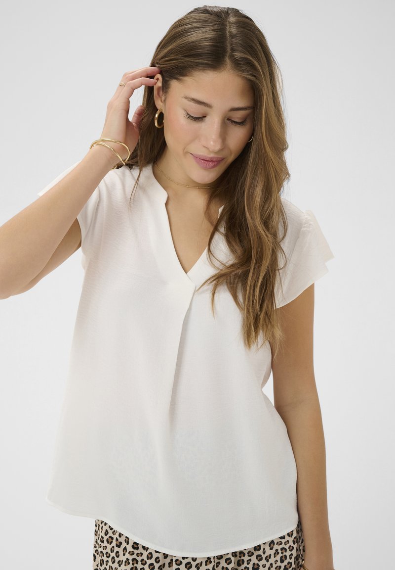 Femme aux longs cheveux bruns portant un chemisier blanc à col en V, des boucles d'oreilles anneaux dorés, des bracelets et un pantalon imprimé léopard, regardant vers le bas et touchant ses cheveux.
