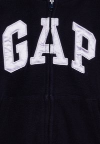Cerca de um close-up de uma jacket de polar azul escuro com letras GAP bordadas em branco e um fecho preto central.