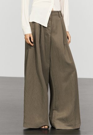 Personne portant un pantalon large vert olive et une chemise blanche à manches longues avec des sandales noires à bouts ouverts sur un sol gris.