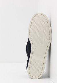 Sohle eines schwarzen Sneakers, der an einer weißen Ecke lehnt und die Größe 42 sowie "HILFIGER" auf der beigen Sohle eingeprägt zeigt.