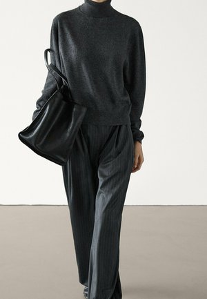 Personne portant un pull à col roulé gris foncé et un pantalon à fines rayures, portant un grand sac cabas en cuir noir.