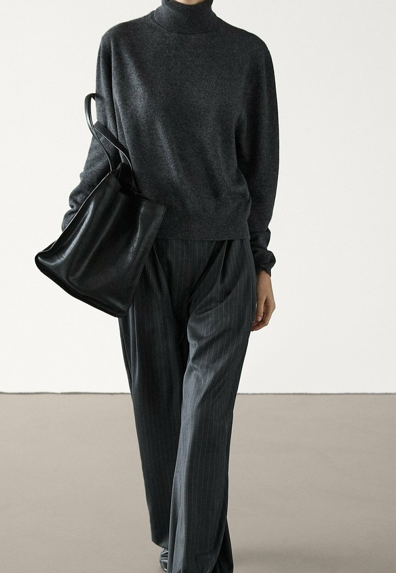 Personne portant un pull à col roulé gris foncé et un pantalon à fines rayures, portant un grand sac cabas en cuir noir.
