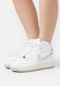 Des jambes portant des baskets Nike montantes blanches avec un swoosh gris clair et des semelles beiges, se tenant sur une surface blanche.