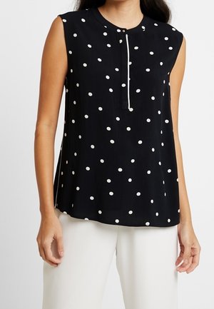 Ärmellose schwarze Bluse mit weißen Punkten, rundem Ausschnitt mit Knopfleiste, getragen mit weißen Hosen.