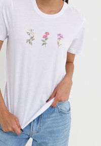 Camiseta de algodón blanca de manga corta, con diseños florales bordados en rosa, púrpura y verde en el pecho.