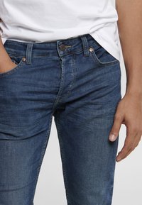 Tmavě modré džíny z denimu s mírným vyblednutím, pět kapes, zipový zesilovač a měděné nýty. Hladká textura s viditelnými detaily švů.