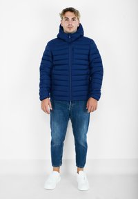 Giacca a vento navy con design a pozzetto orizzontale, chiusura con zip e cappuccio, abbinata a jeans azzurri e sneaker bianche.