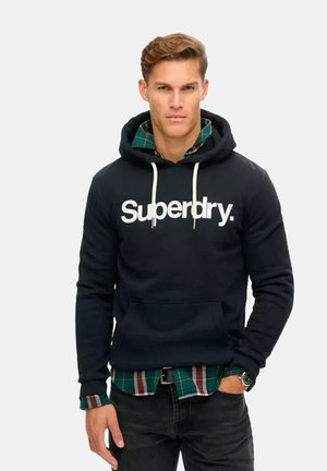 Haut à capuche noir avec logo "Superdry" blanc, capuche avec cordon de serrage, poche kangourou. Superposé sur une chemise à carreaux verte, avec un jean noir et une montre.