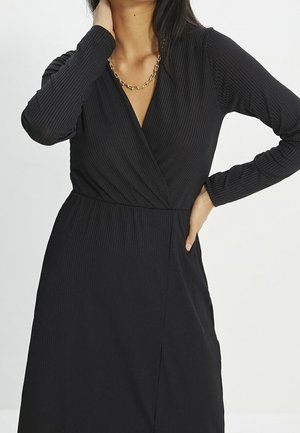 Frau trägt ein langärmeliges schwarzes Rippenkleid und eine goldene Halskette, die eine Hand am Hals und die andere an der Hüfte hat.