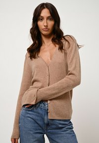 Hellbraunes Kaschmir-Cardigan mit V-Ausschnitt, Knopfleiste und langen Ärmeln. Weicher Griff und leichter Stretch, kombiniert mit blauen Denim-Jeans.