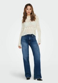 Pull en tricot crème avec motif ajouré et ourlet court, associé à un jean flare taille haute bleu foncé. Des bottines noires viennent compléter la tenue.