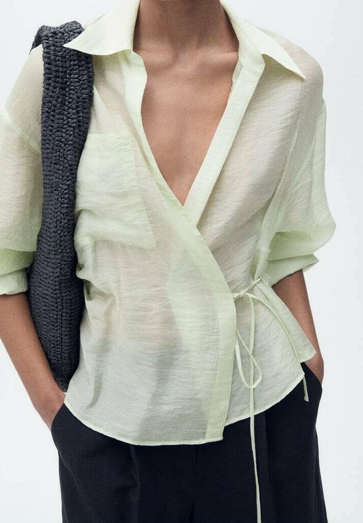 Personne portant un chemisier cache-cœur vert clair semi-transparent avec ceinture à la taille, pantalon noir, et portant un sac à épaule texturé gris foncé.