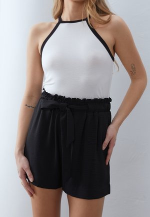 Vrouw draagt een mouwloze witte haltertop met zwarte bies en een zwarte short met hoge taille die met een riem is vastgestrikt, staand tegen een effen achtergrond.