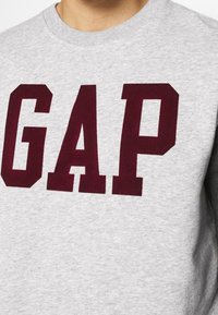 Šedá mikina s výrazným vínovým nápisem "GAP", ribbovaným výstřihem a měkkou texturou. Detailní pohled na přední design.