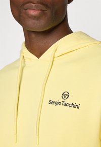 Gul bomulds hoodie med snøre, ribbede kanter og et sort broderet logo. Stoffet har en glat tekstur.