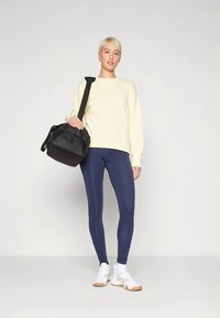 Persona in piedi che indossa una felpa gialla chiaro, leggings blu navy, sneakers bianche e tiene in mano una borsa a spalla nera.