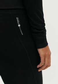 Zwarte langemouwen top gemaakt van zachte stof, met een getailleerd ontwerp en een geborduurd logo aan de zijkant. Gladde textuur en subtiele naden.