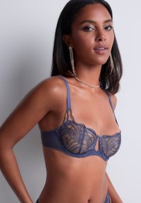 Soutien-gorge en dentelle bleu marine avec tissu transparent, support à armatures et accents dorés décoratifs sur les bretelles. Broderie complexe et détail découpé.