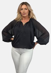 Czarna bluzka w kropki z długimi bufkami, zebranym dekoltem i subtelnym blaskiem; zestawiona z białymi dopasowanymi spodniami dla kontrastu.