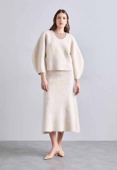 Lys beige strikket sweater med ballonærmer, parret med en matchende knælang nederdel. Begge beklædningsgenstande har en blød, fyldig tekstur.