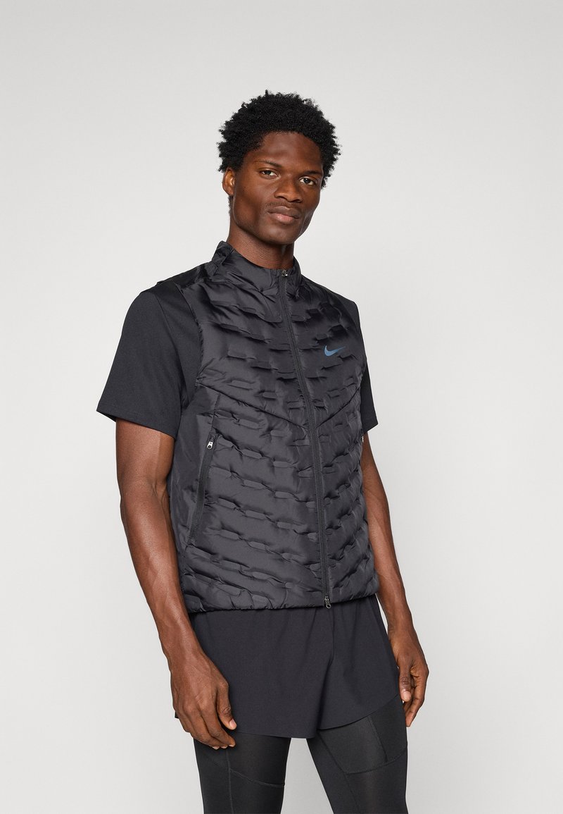 Vesta puffer neagră cu o suprafață texturată și logo-ul Nike albastru, asociată cu un tricou negru cu mânecă scurtă și pantaloni scurți, stând pe un fundal neutră.