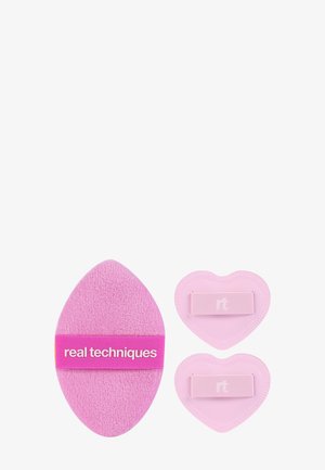Lyserød Real Techniques makeup-applikator med to mindre lyserøde hjerteformede puder, alle med stofgreb på en hvid baggrund.