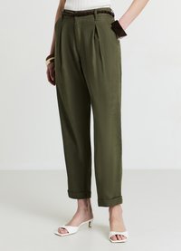 Pantalon vert olive, avec des plis sur le devant et des revers roulés. Confectionné dans un tissu lisse, associé à des talons blancs à bout ouvert.