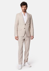 Jasno beżowy garnitur o teksturowanym wykończeniu, z kołnierzem z wycięciem, dwoma guzami, spodniami o prostym kroju oraz zestawiony z białymi trampkami.