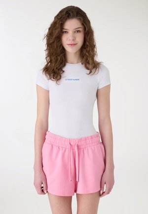 Giovane donna con capelli ricci che indossa una t-shirt bianca aderente con scritte blu e pantaloncini rosa con coulisse, su uno sfondo semplice.