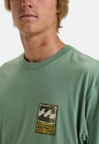 Triko s krátkým rukávem v mátově zelené barvě má na levé straně hrudi výraznou nášivku s logem v žluté a černé barvě. Měkký bavlněný materiál.